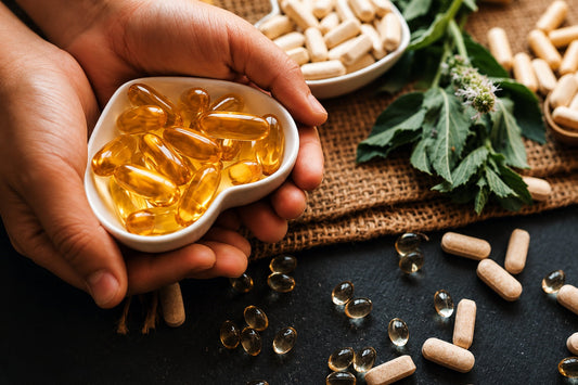 Hands holding a heart shaped dish of omega-3 capsules beside supplement capsules and fresh herbs